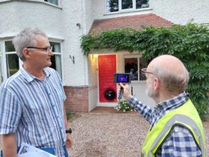 Picture of Louis Stephen with Brian Harper from Energy Tracers. Louis is looking at the thermal imaging camera pointing at house. In background you can see a large fan temporarily installed at the front door.